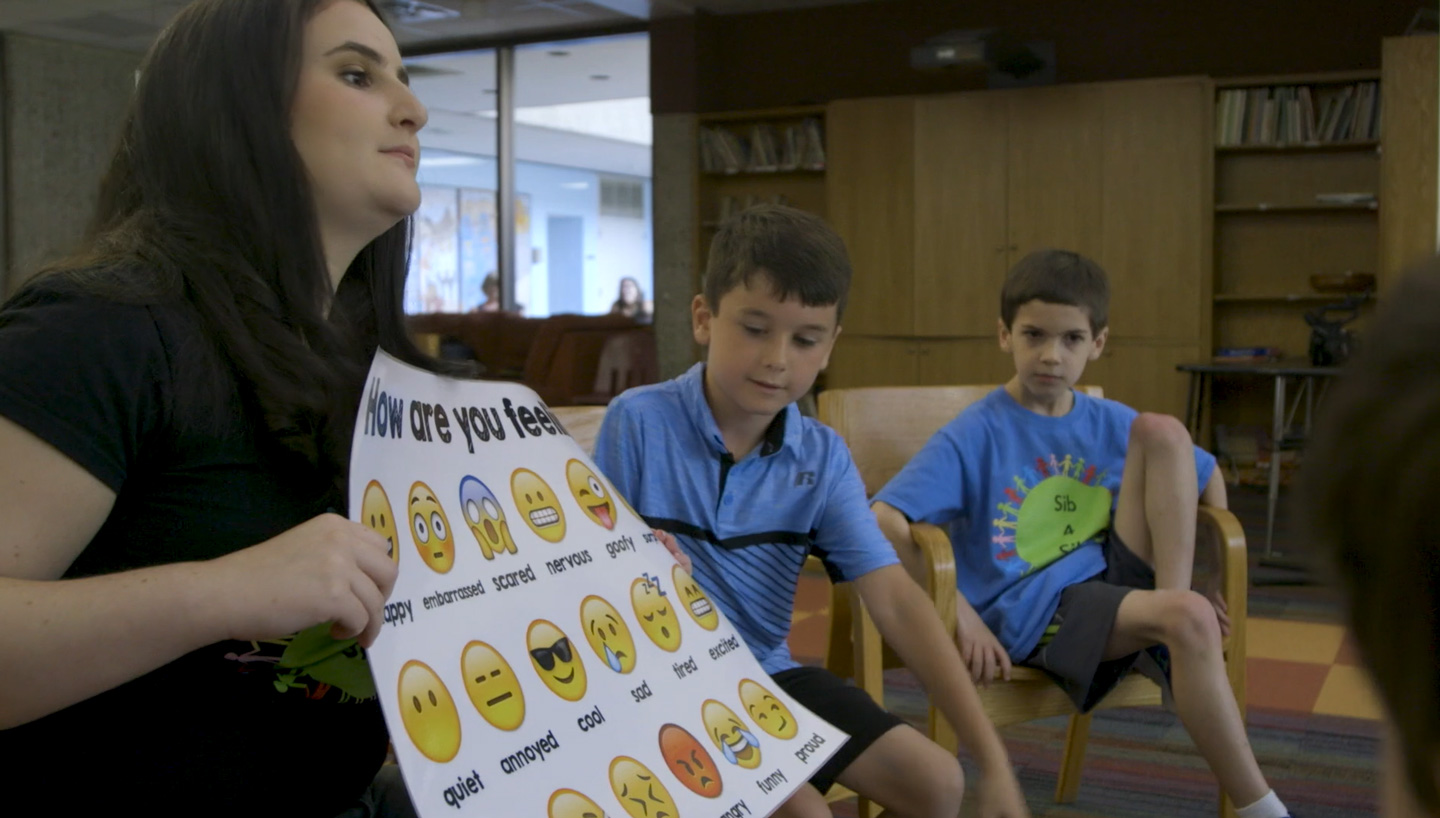 Jessica holding a feelings poster of emojis while 2 children look on.
