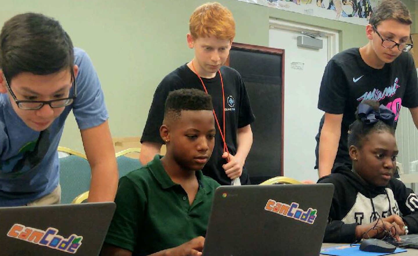 Noah and two other teens looking over the shoulders of young students at laptop computers.
