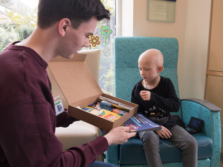 Malcolm sits in a hospital room with a young patient showing him a box of art supplies.