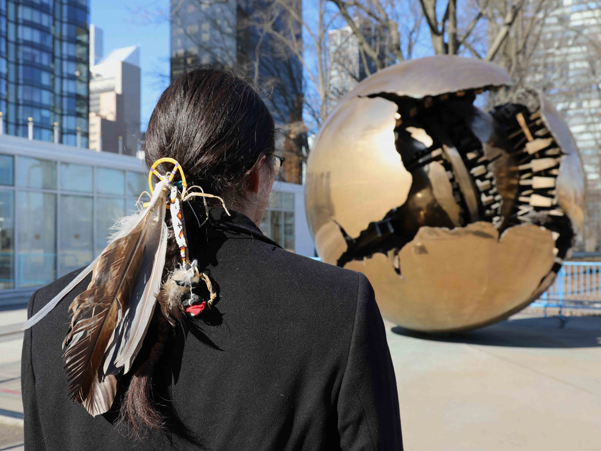 Nathan is standing outside the United Nations building facing a sculpture with his back to the camera. His long hair is tied back in a ponytail with Native American insignia and feathers attached to it.