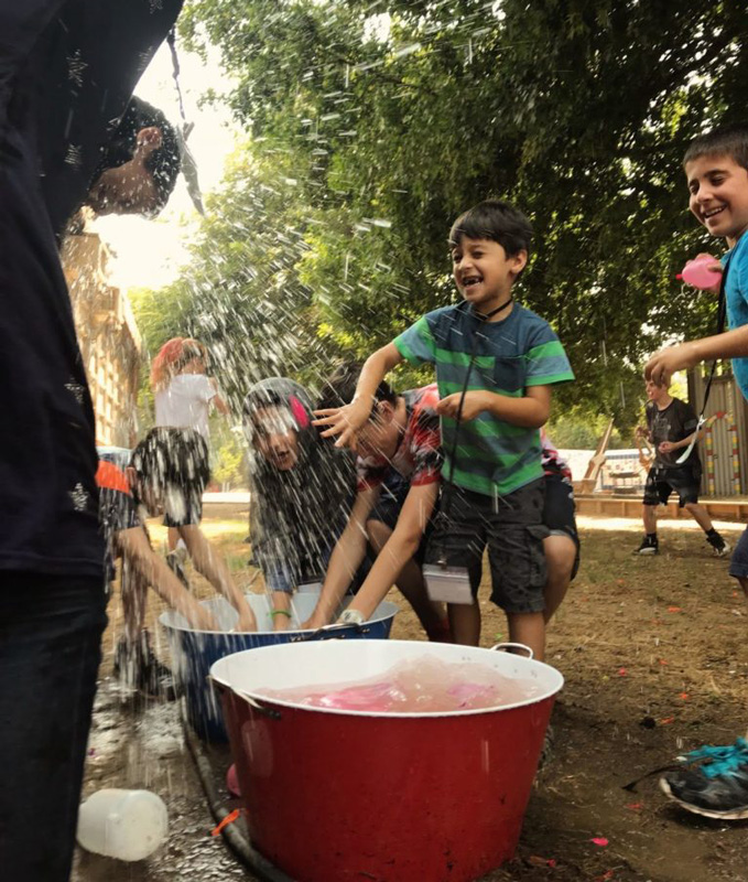 Children laughing and playing with water in large tubs at camp while more water is sprayed on them.