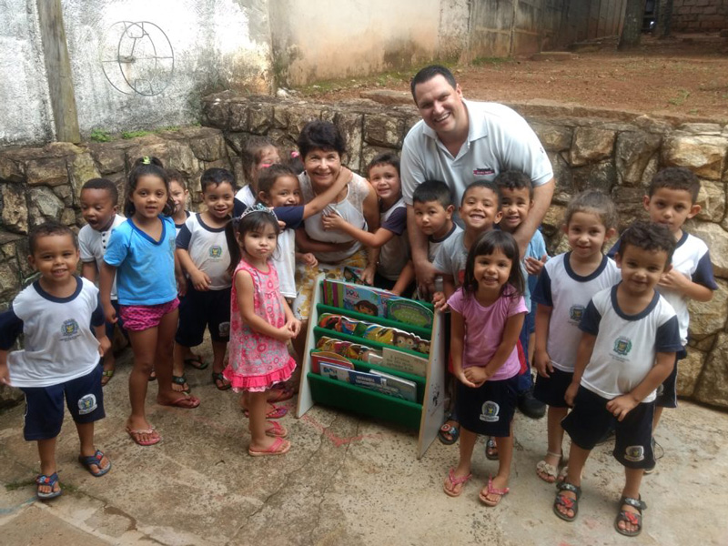 Many young children and 2 adults outside a Brazilian daycare standing by a small display case of books.
