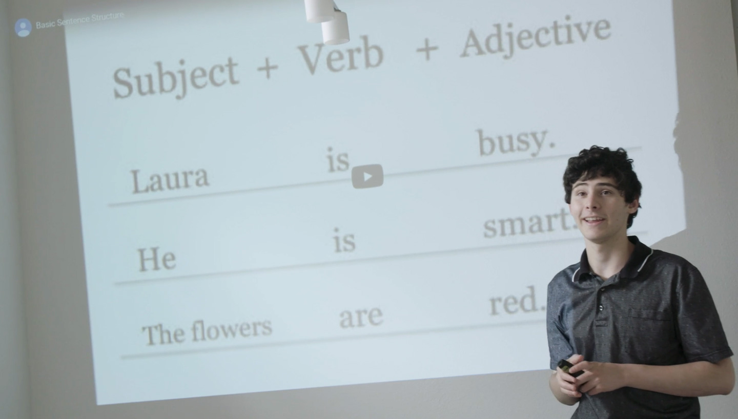 John standing at the front of a classroom with a language lesson displaying on the large projector screen.