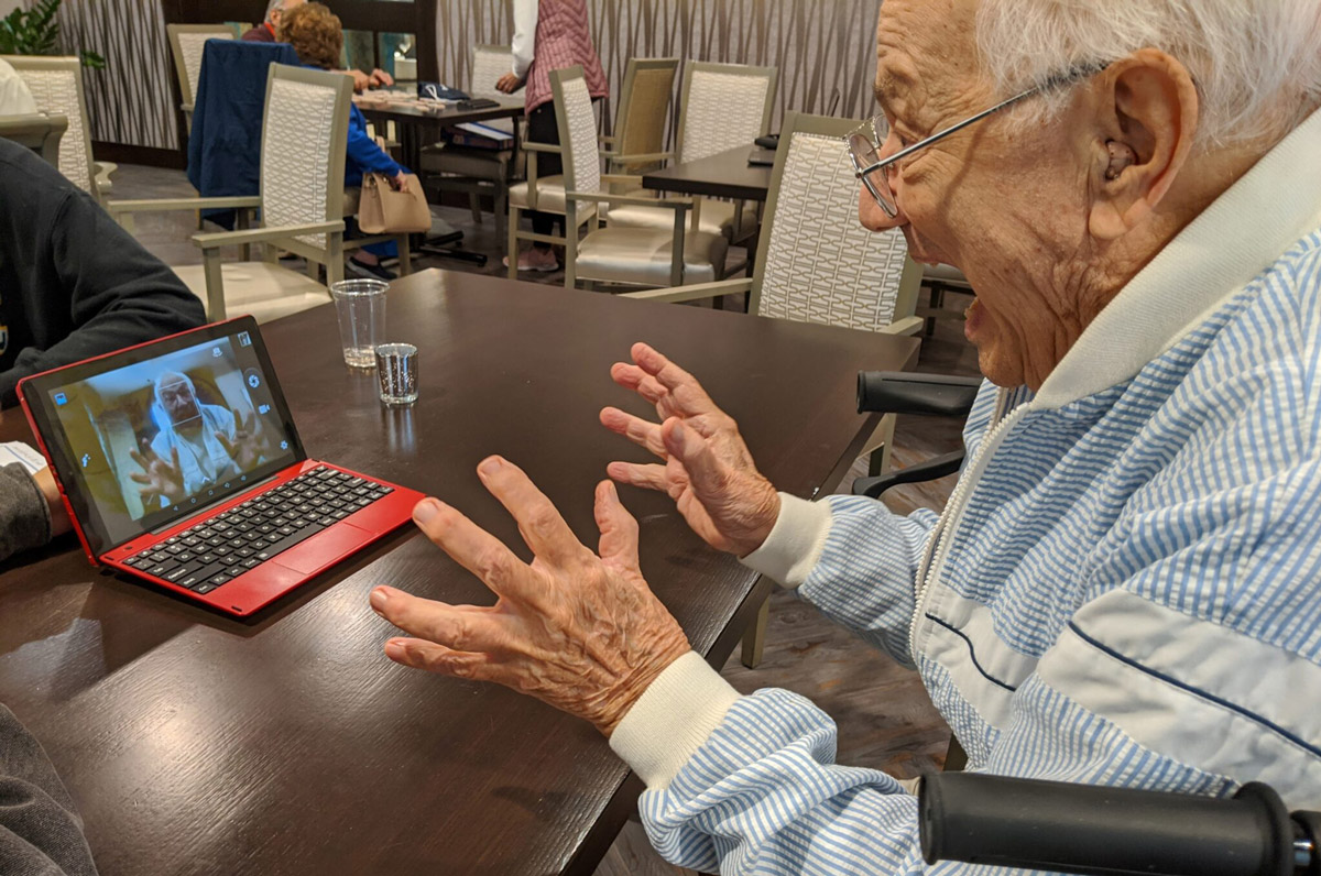 An elderly man sits in front of a laptop with its camera on and he expresses surprise at the image of himself on the screen.