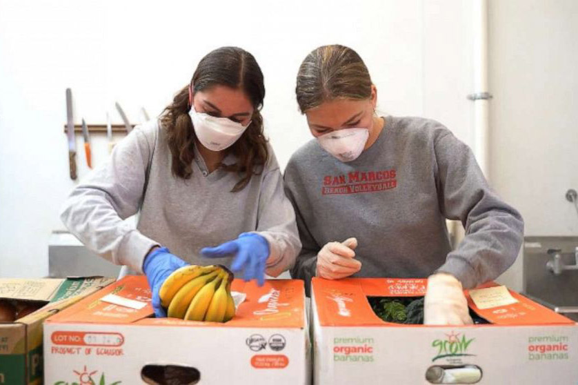 Two teenage girls wearing masks pack large food boxes with fruit and vegetables.