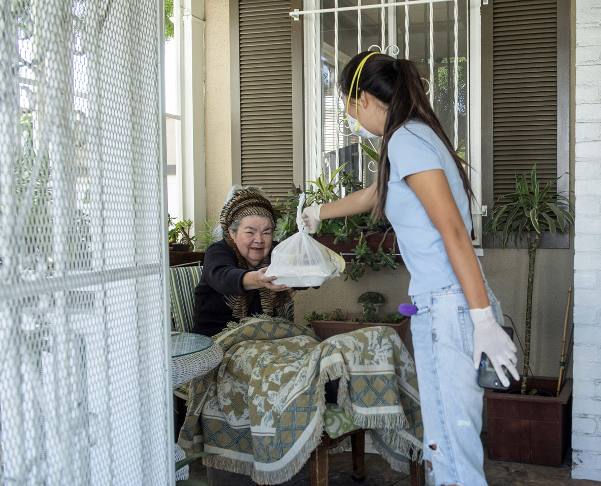 A teen wearing a mask and gloves hands a prepared meal in a plastic bag to an elderly woman in her home.