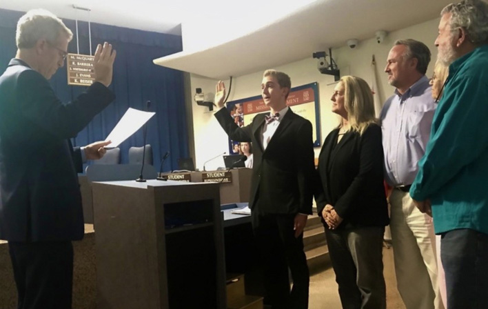 Zachary stands with his right hand in the air taking an oath administered by another adult with his right hand in the air and reading a script.