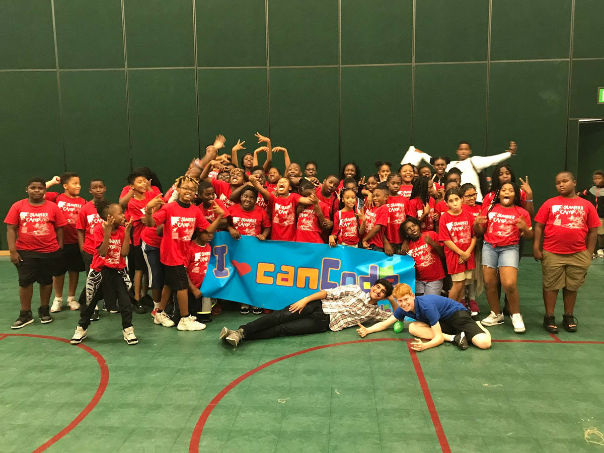 Rubin poses in front of a large group of about 30 elementary age children wearing red camp tshirts. The children are holding a banner that says "I heart can code".