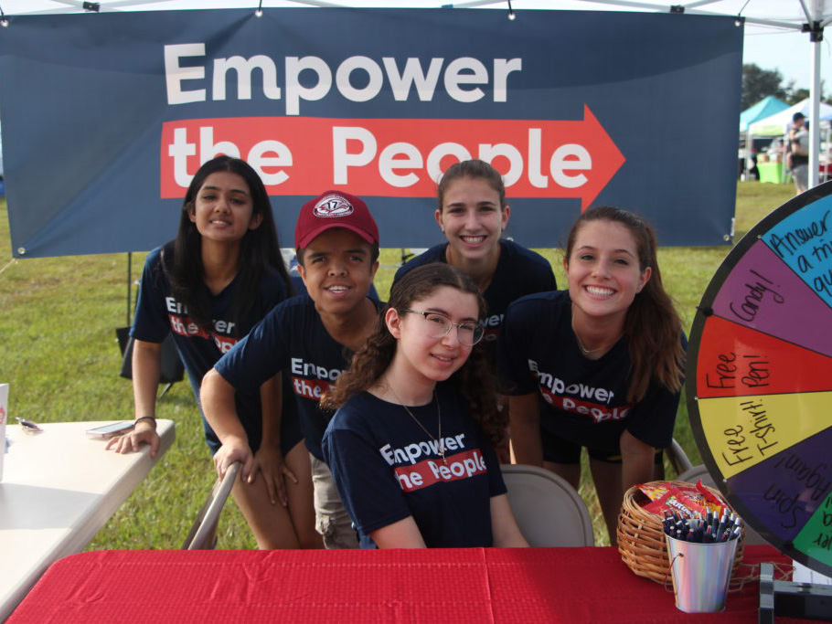 Casey and 4 other teens at an outdoor booth with the organization's logo banner behind them. The booth table has pens, candy, and a prize wheel for spinning.