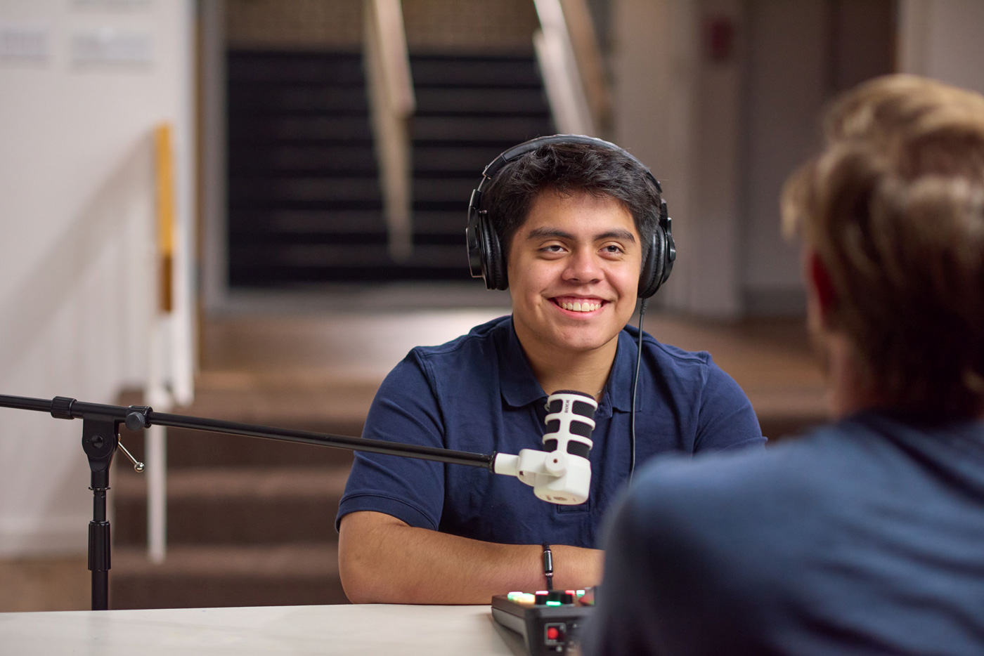 Two people recording a podcast, one wearing headphones, speaking into a mic at a table with audio equipment.