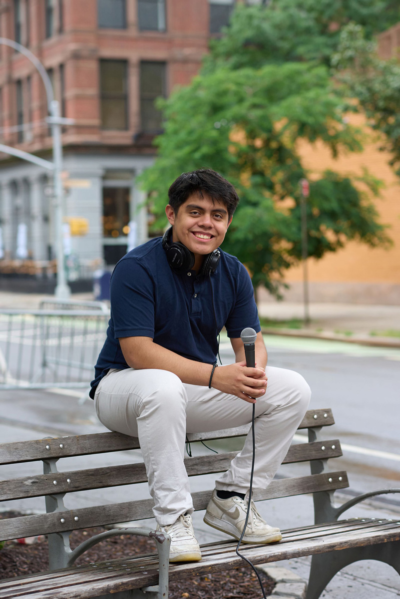 Person sitting on a bench outdoors, holding a microphone and wearing headphones around their neck, with a street scene behind.