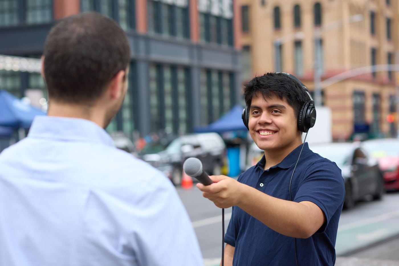 Outdoor interview scene with one person speaking into a microphone and wearing headphones, facing another person in a light blue shirt.