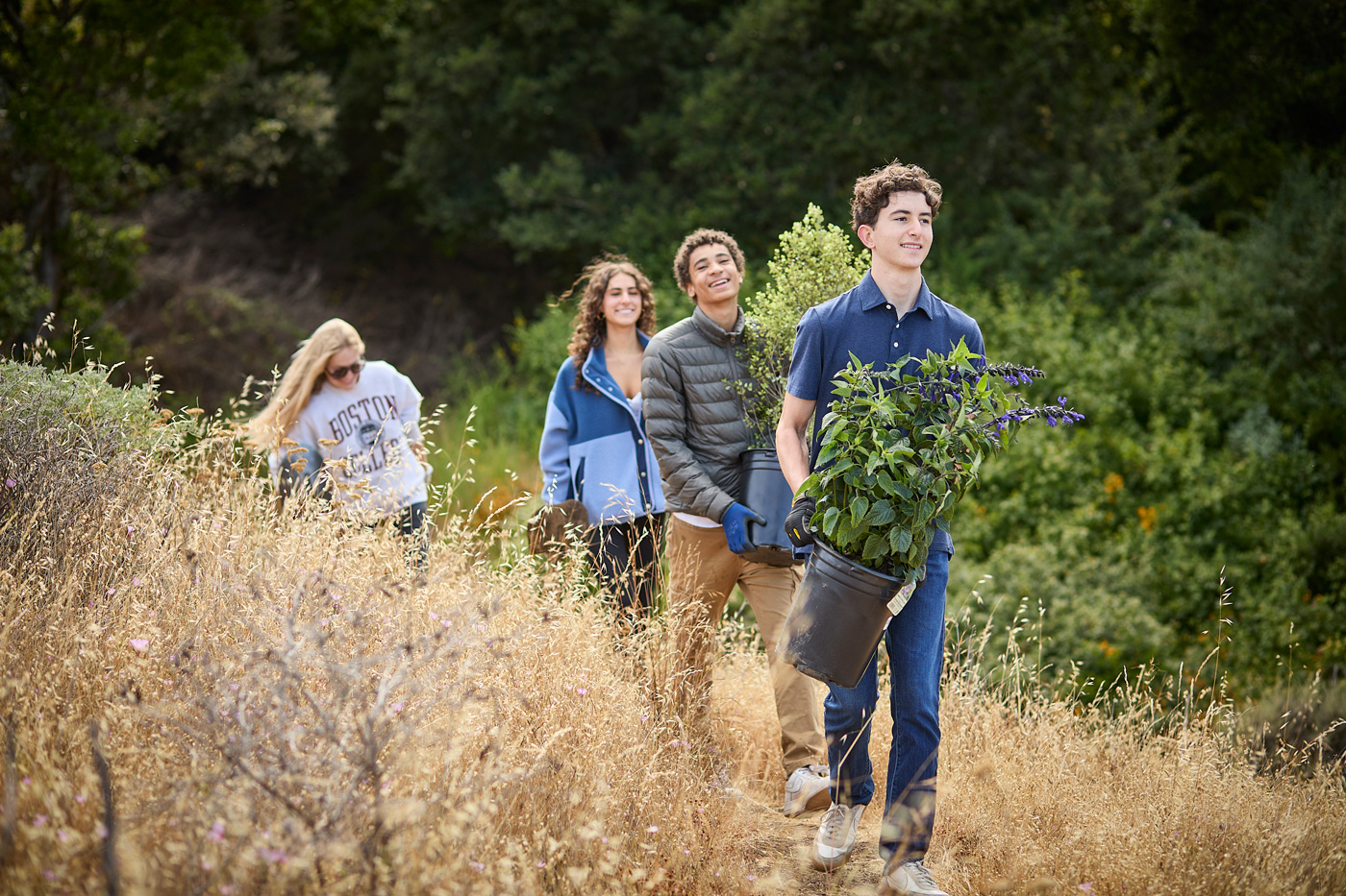 Four people walking a trail in nature; the lead carries a potted plant with purple flowers, others follow with casual outdoor gear.