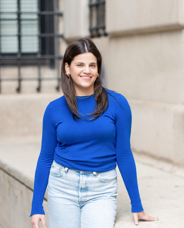 Bella standing casually in front of a beige building, wearing a bright blue shirt and jeans, with a stone bench and window grilles behind.
