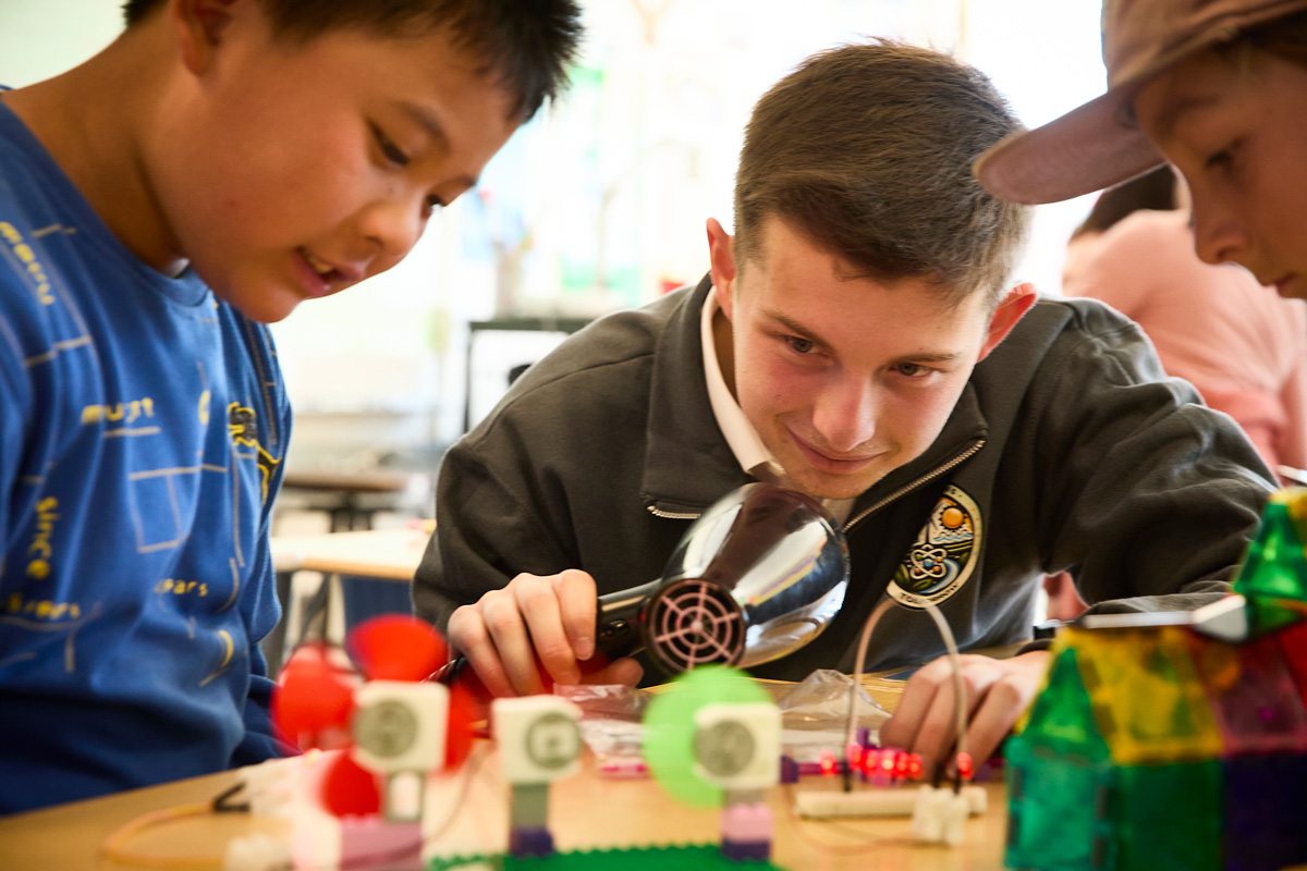 Three people collaborating on a STEM project indoors, using fans, wires, and plastic components, with one holding a hairdryer for testing.