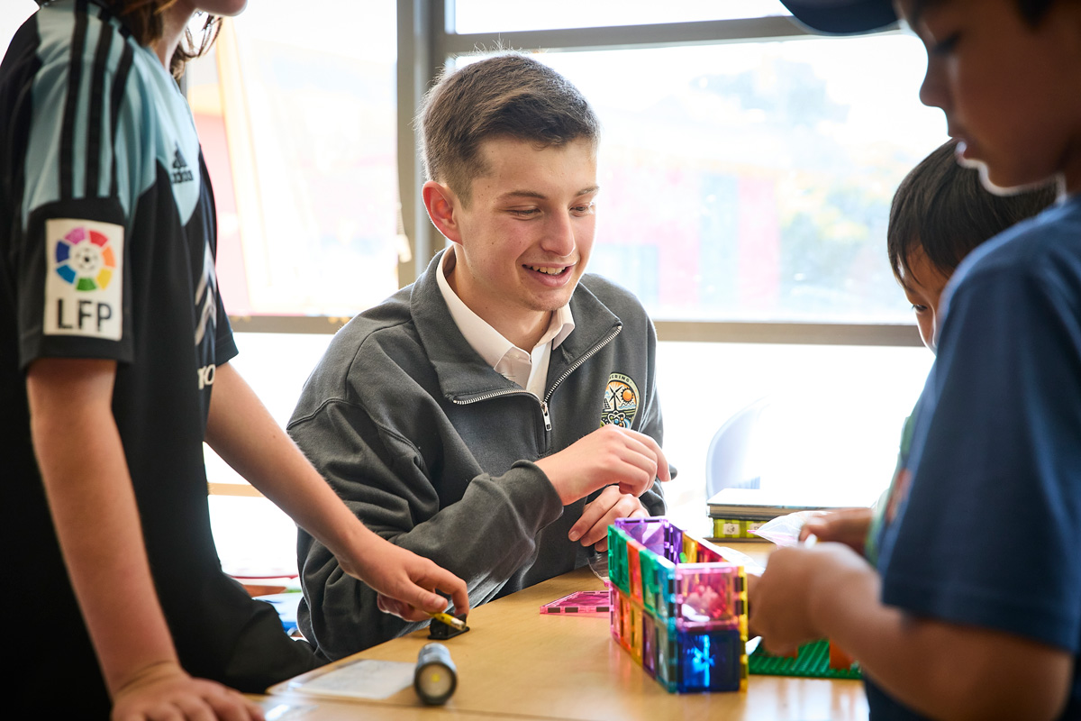 Teenage student smiling while helping younger children build a colorful structure with magnetic tiles at a classroom table.