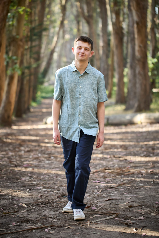 Ari walking on a forest path, wearing a short-sleeve button-up shirt and navy pants, smiling with trees in the background.