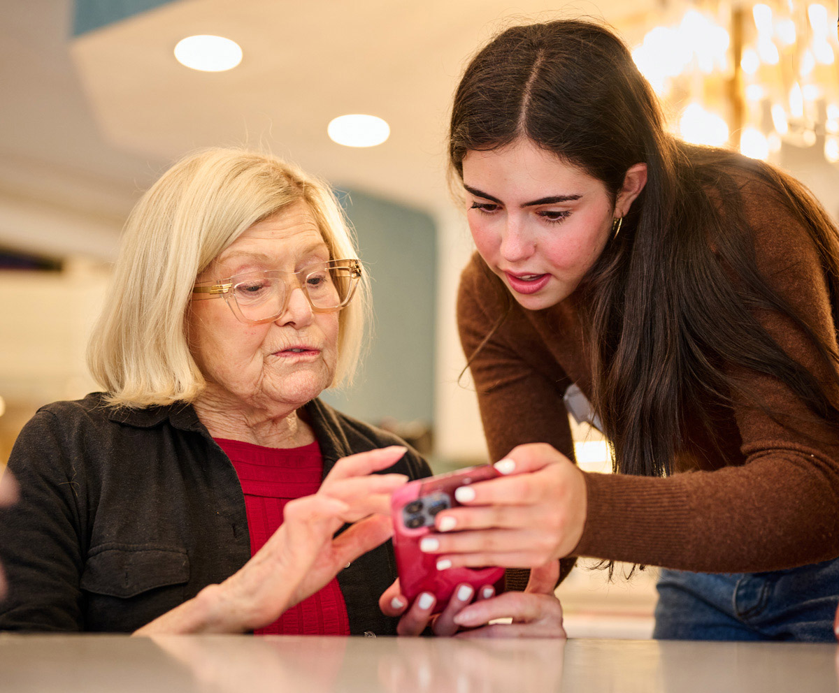 Two people looking at a smartphone together indoors—one seated and holding the phone, the other standing and assisting with a gesture.