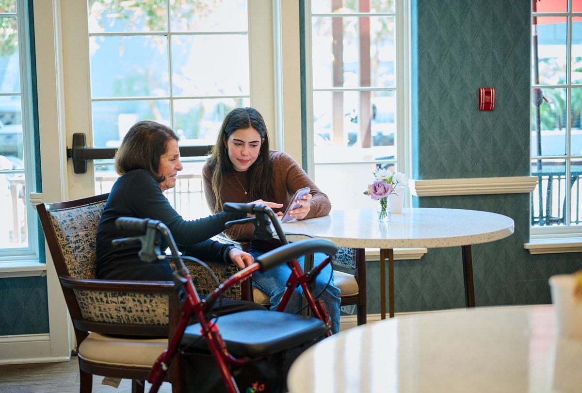 Two people at a round table near large windows, one showing a smartphone to the other beside a red walker and a vase of flowers.