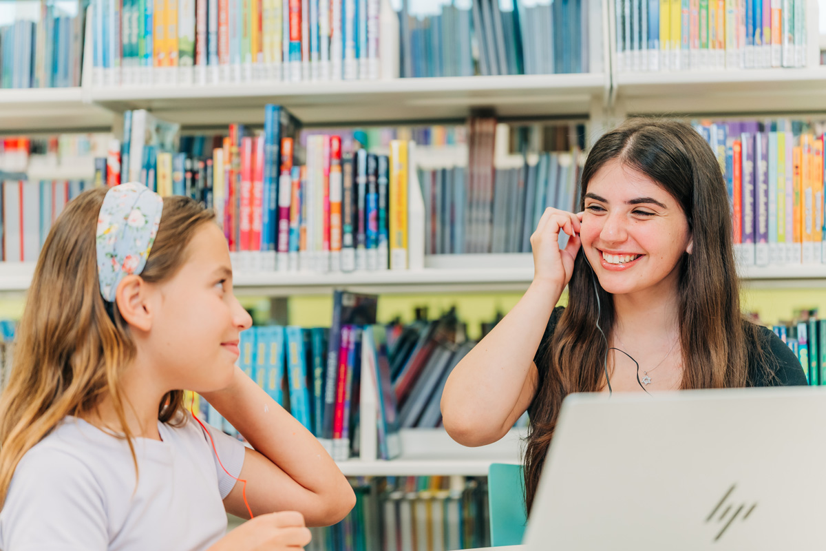 Two people sitting in a library with bookshelves behind them, both wearing earphones; one uses an HP laptop, the other wears a floral headband.