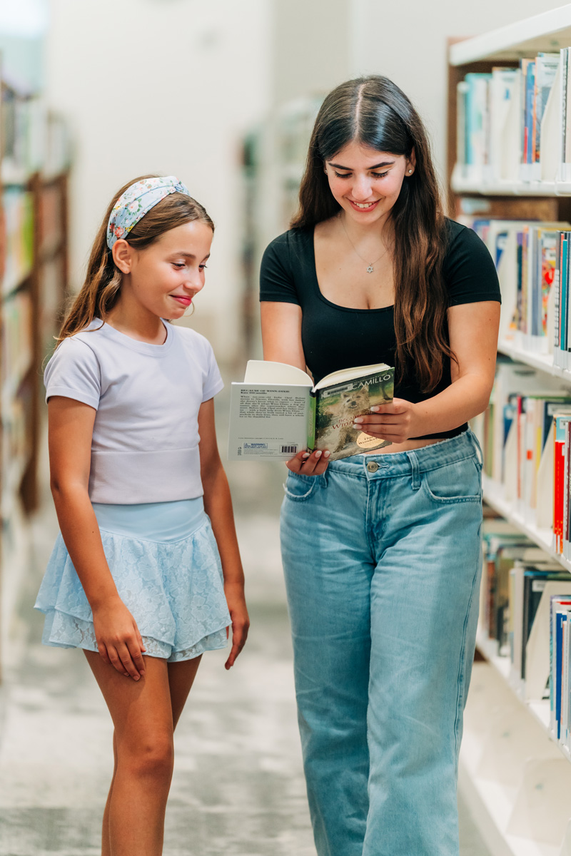 Two people standing in a library, one holding and showing a book to the other, surrounded by tall shelves filled with books.