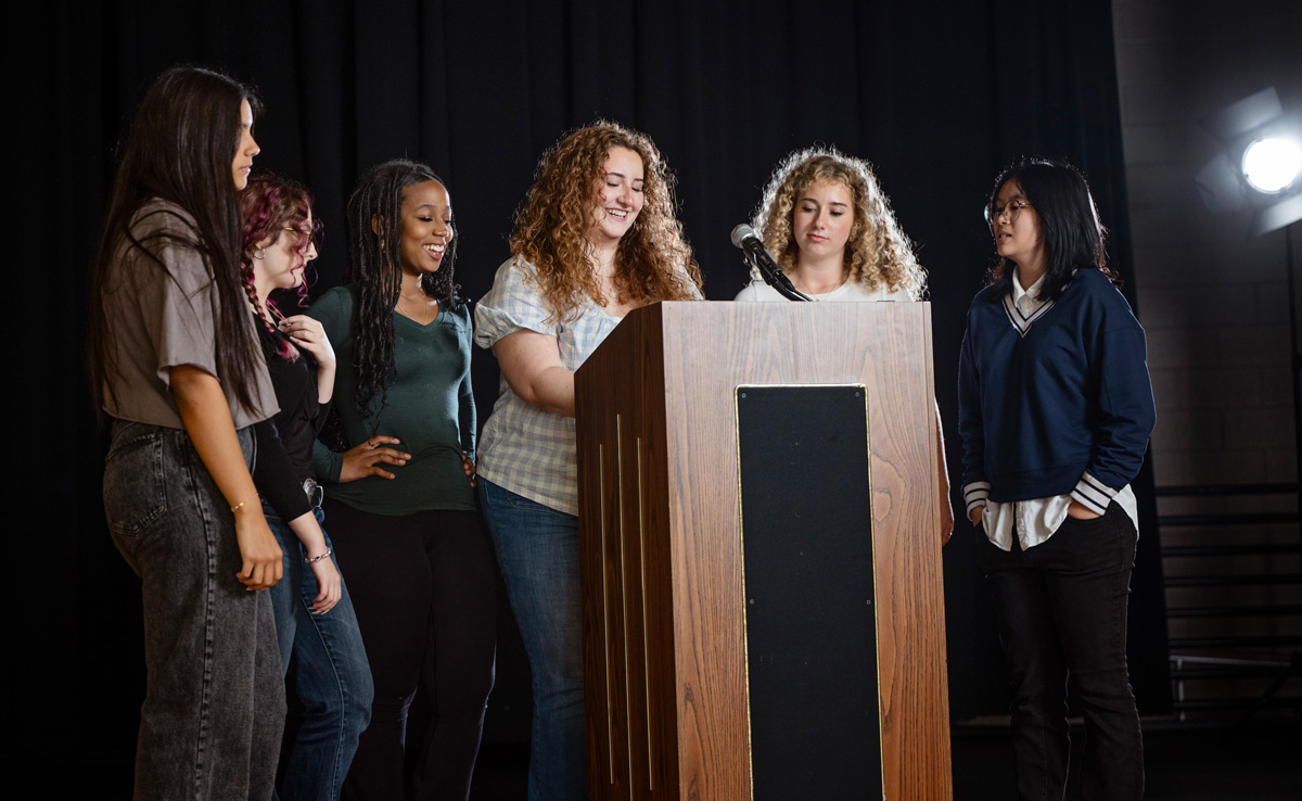 Six people gathered around a wooden podium with a microphone, lit dramatically against a dark background, suggesting a group presentation.