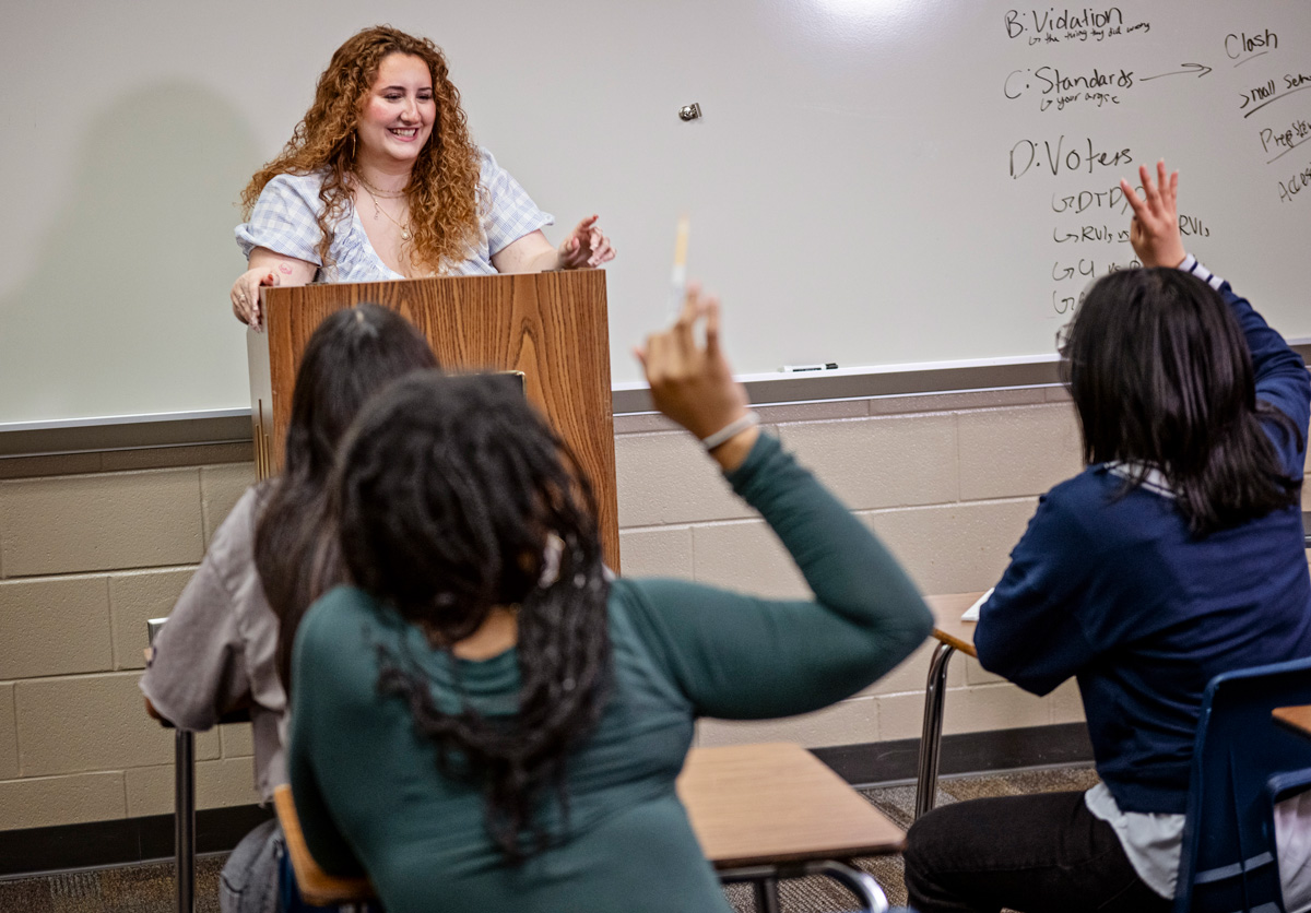 Classroom scene with a speaker at a wooden podium, students seated at desks, and two raising hands near a whiteboard with notes.