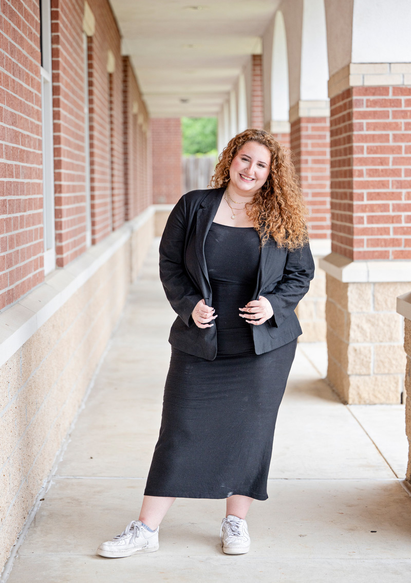 Alexandria standing in a brick-arched corridor, wearing a black blazer and dress with white sneakers, hands holding blazer, greenery behind.