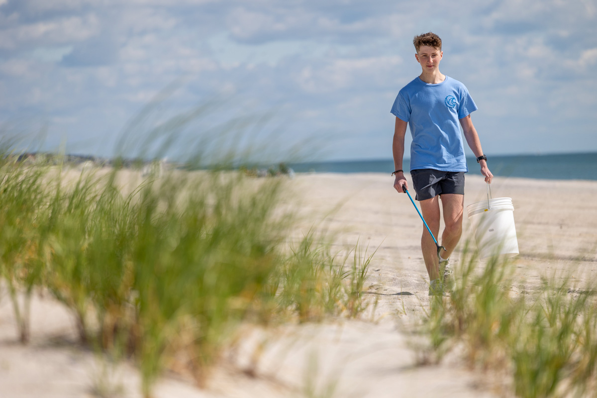 Landon walking barefoot on a sandy beach, wearing a blue T-shirt and dark shorts, carrying a white bucket and a blue-handled tool.