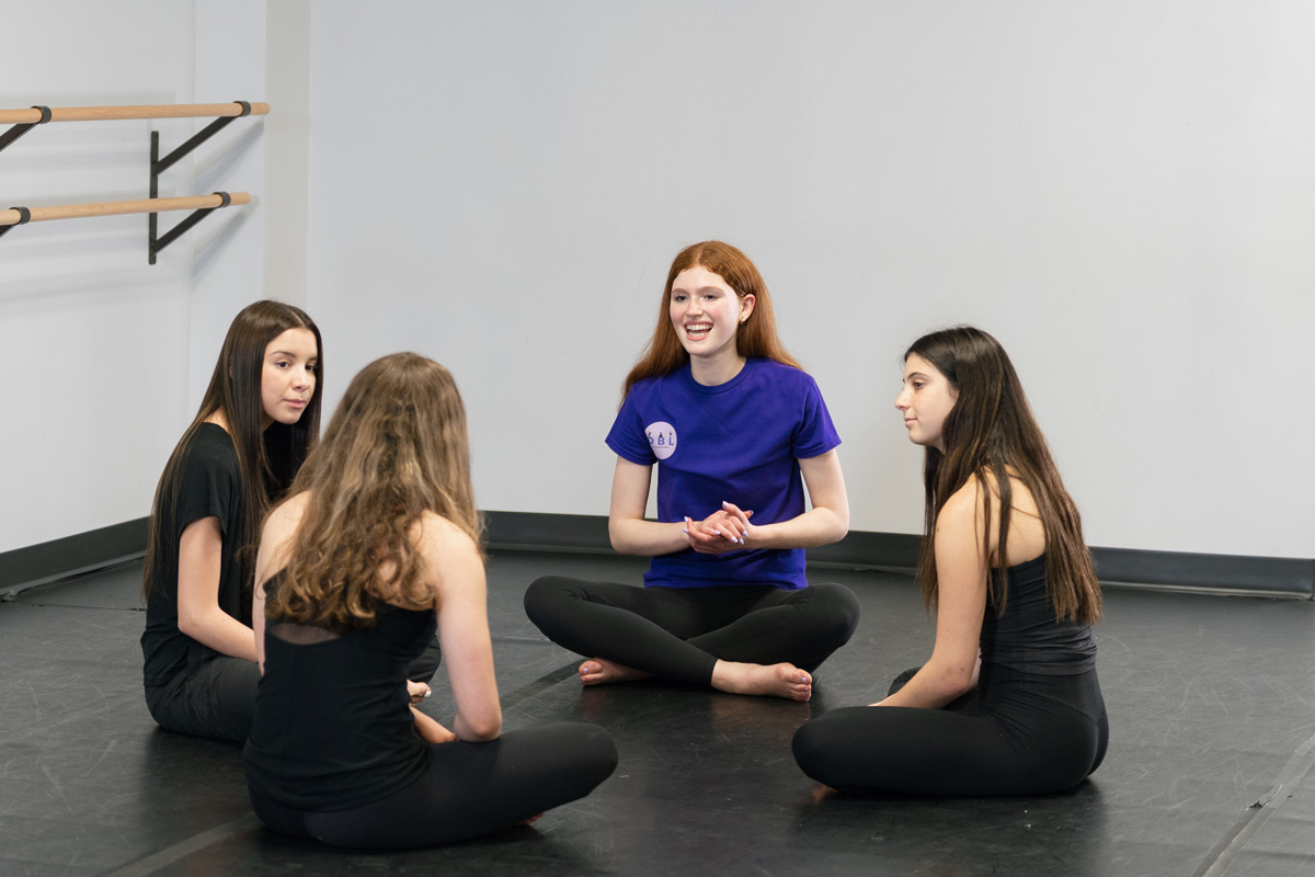 Individuals seated in a circle on the floor of a dance studio, near ballet barres, engaged in conversation; one wears a blue logo shirt.