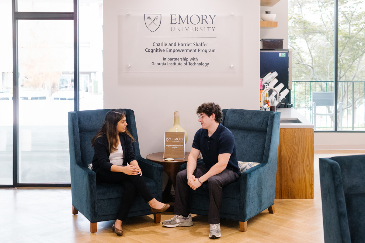 Two people seated in blue armchairs in a sunlit room with a kitchenette, talking beside a plaque honoring Emory’s Cognitive Empowerment Program.
