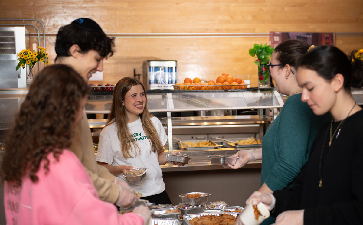 People packing up food in a cafeteria-style setup with trays of fried items and pasta; one wears a “FRESH OPPORTUNITIES” shirt near oranges and flowers.