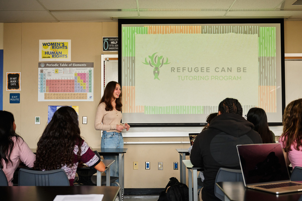 Classroom presentation introducing the “Refugee Can Be Tutoring Program” with students seated, posters on the walls, and a large screen displaying the program’s name and logo.
