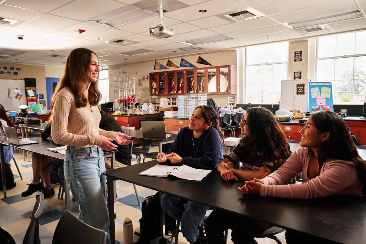 Student standing at the front of a classroom during a lesson, with others seated at desks near flags, posters, and shelves of trophies.