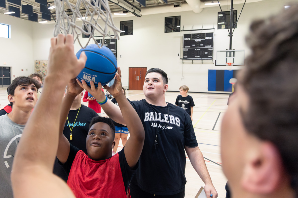 Basketball training session in a gym, where a participant holds a blue “USA Official Size” ball as a coach in a “BALLERS Aid Another” shirt offers guidance amid other team members and equipment.