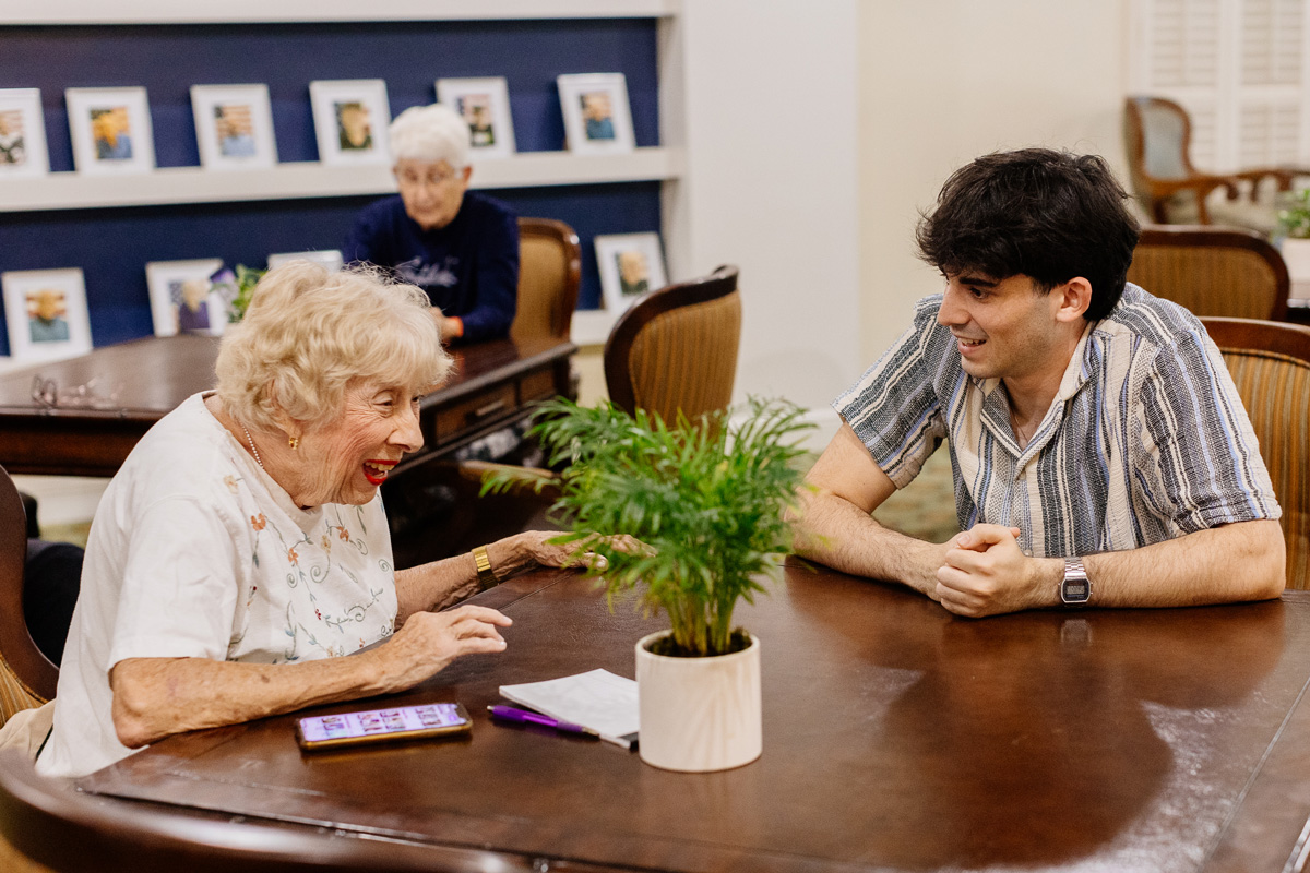 Two people sit talking at a small table with a plant, paper, and phone; another person sits in the background by framed photos.