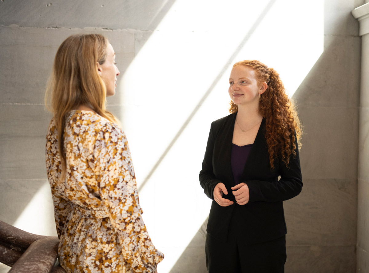 Two people talk indoors as sunlight creates bright diagonal patterns across the wall behind them.