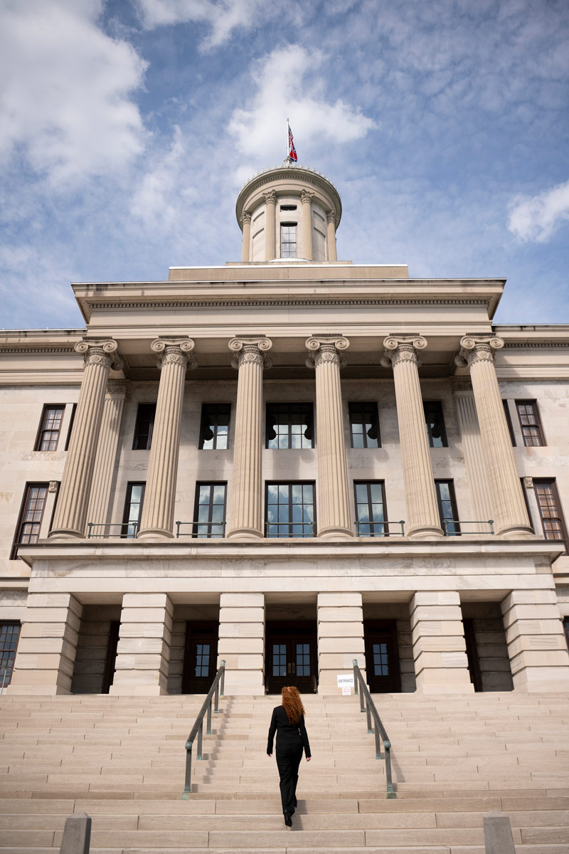 Person walks up wide stone steps toward a large columned government building topped with a dome and flag.