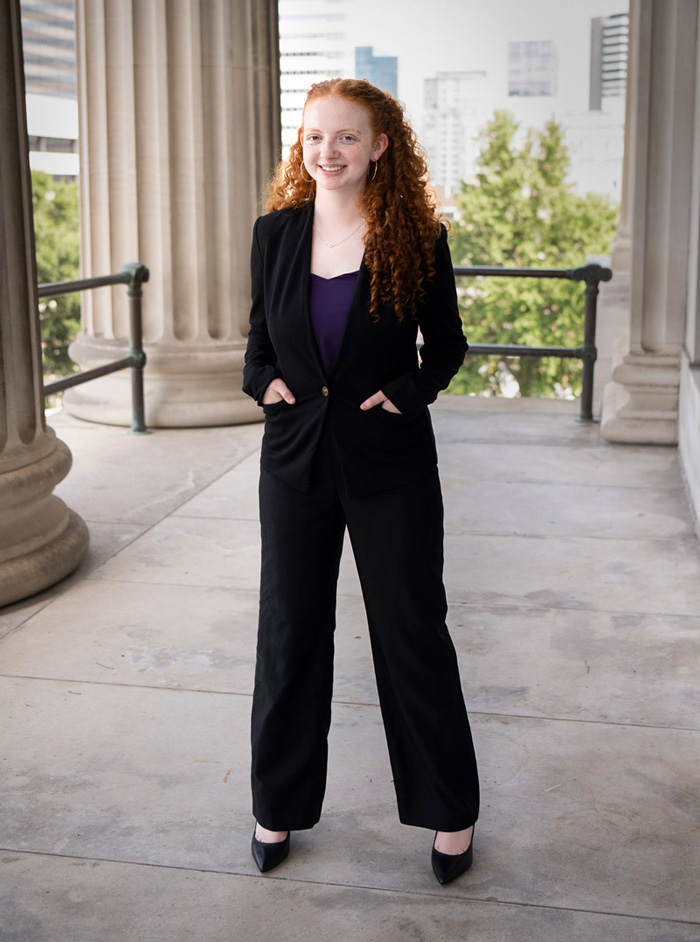 Person stands on a stone terrace between tall columns with a city skyline visible in the background.
