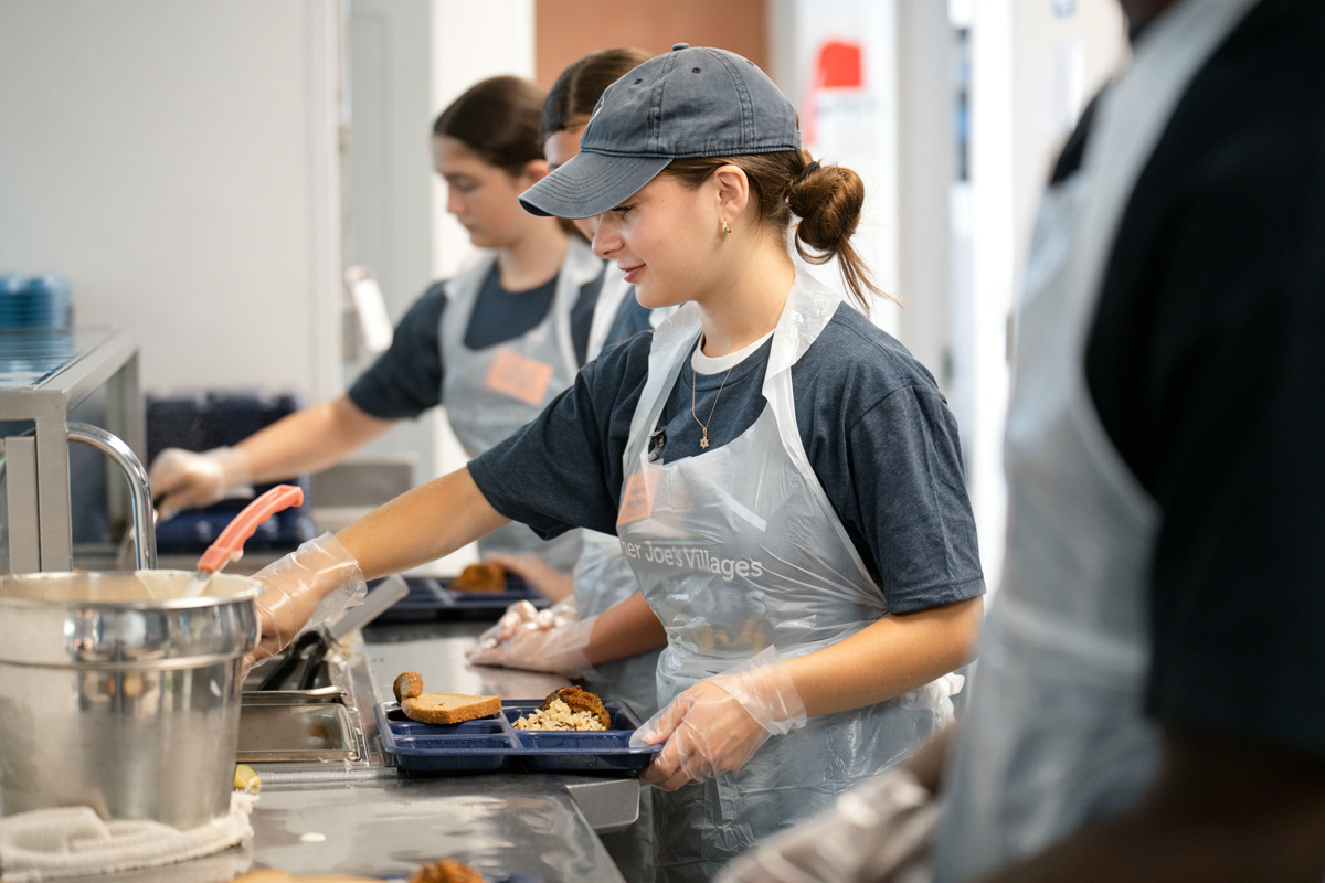 Volunteers in aprons and gloves serve food onto trays in a busy community kitchen.