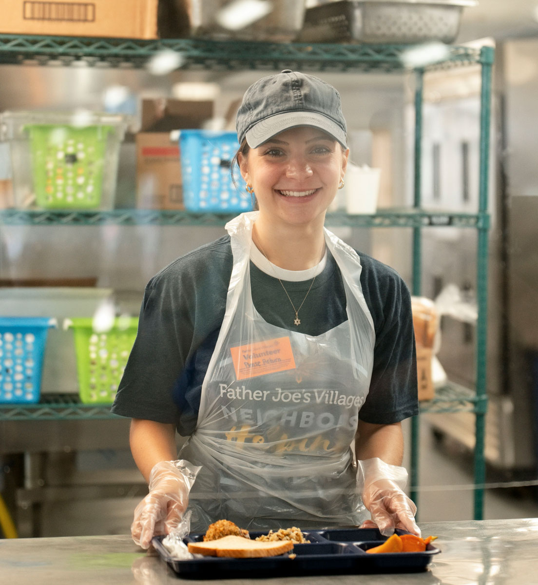 Volunteer in apron and gloves holds a food tray in a kitchen with shelves of supplies behind them.