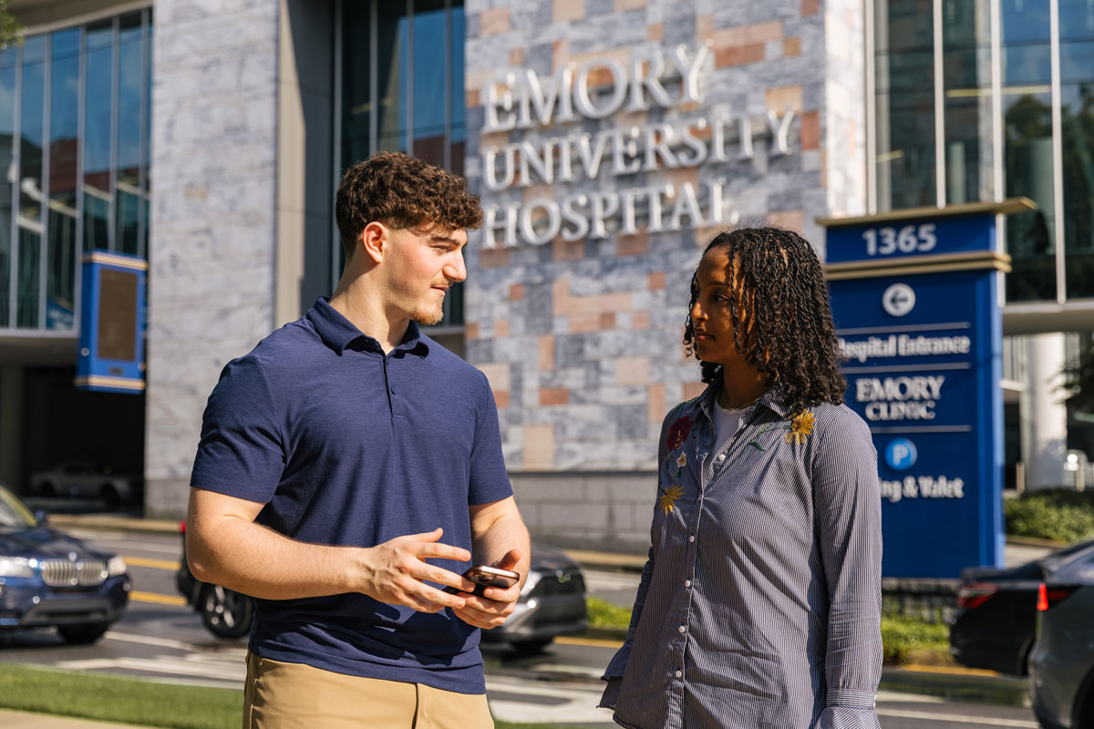 People talk outside Emory University Hospital near entrance signs and parked cars.