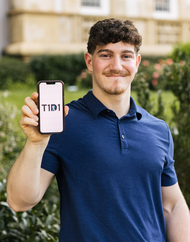 Person holds a smartphone displaying “T1D1” while standing outside near greenery and a building.