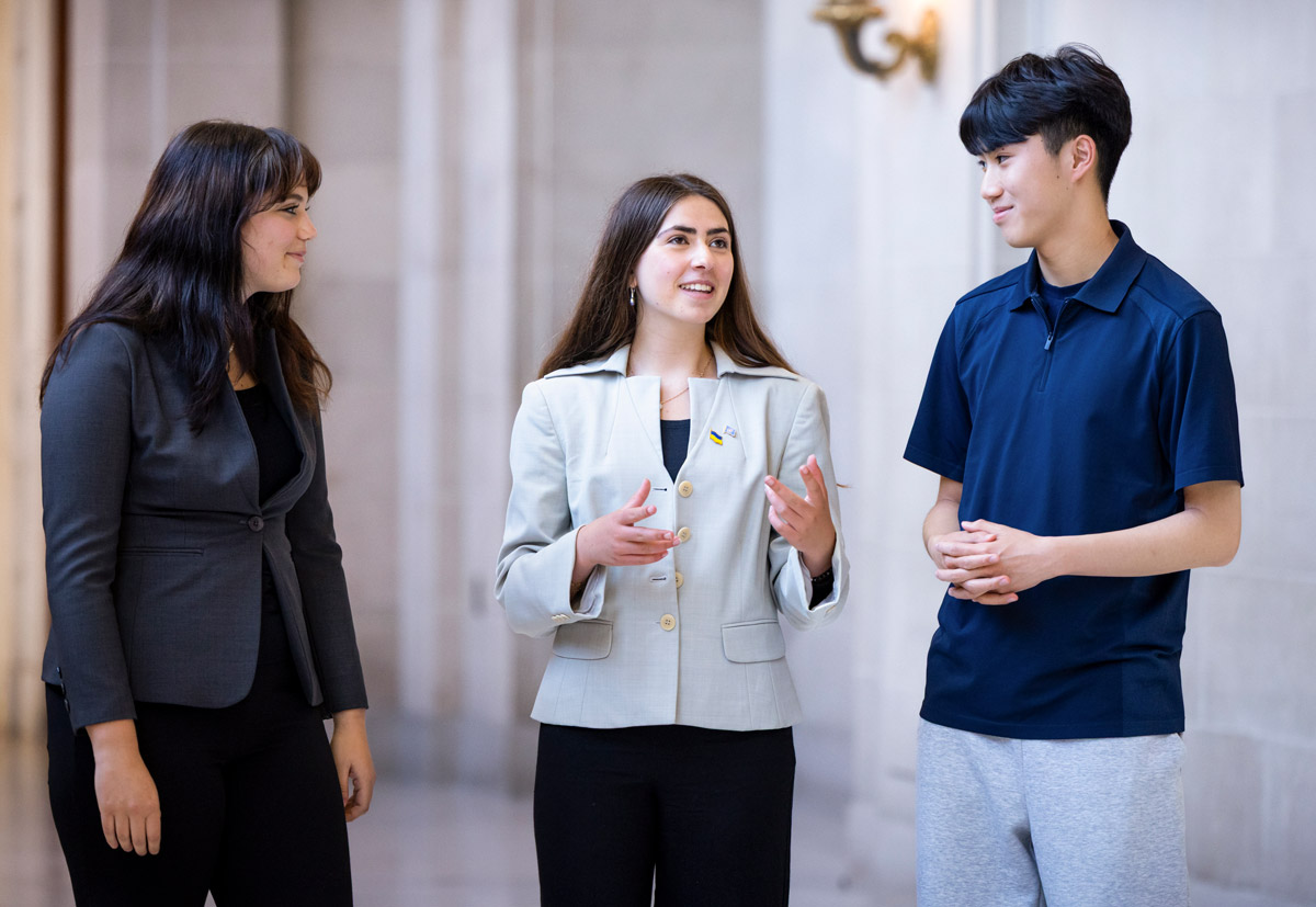 Three people stand in a hallway talking as the person in the center gestures with both hands.