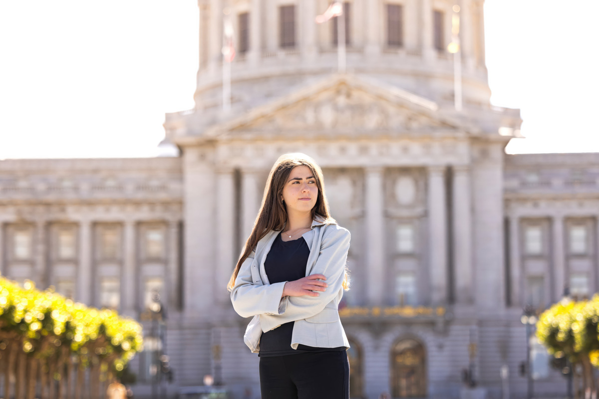 A person stands outside with arms crossed in front of a large domed building lined with columns and trees.