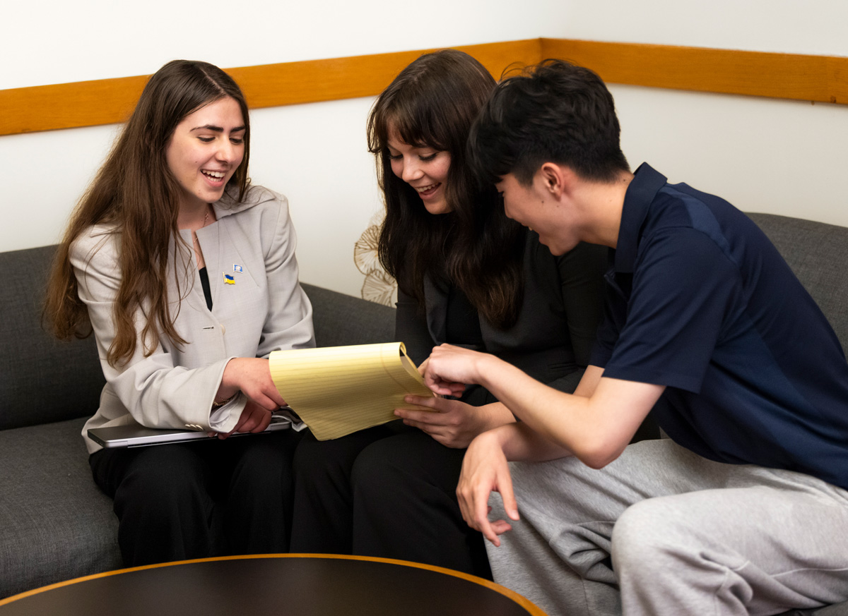 Three people sit on a couch reviewing a yellow notepad together in a well‑lit room.