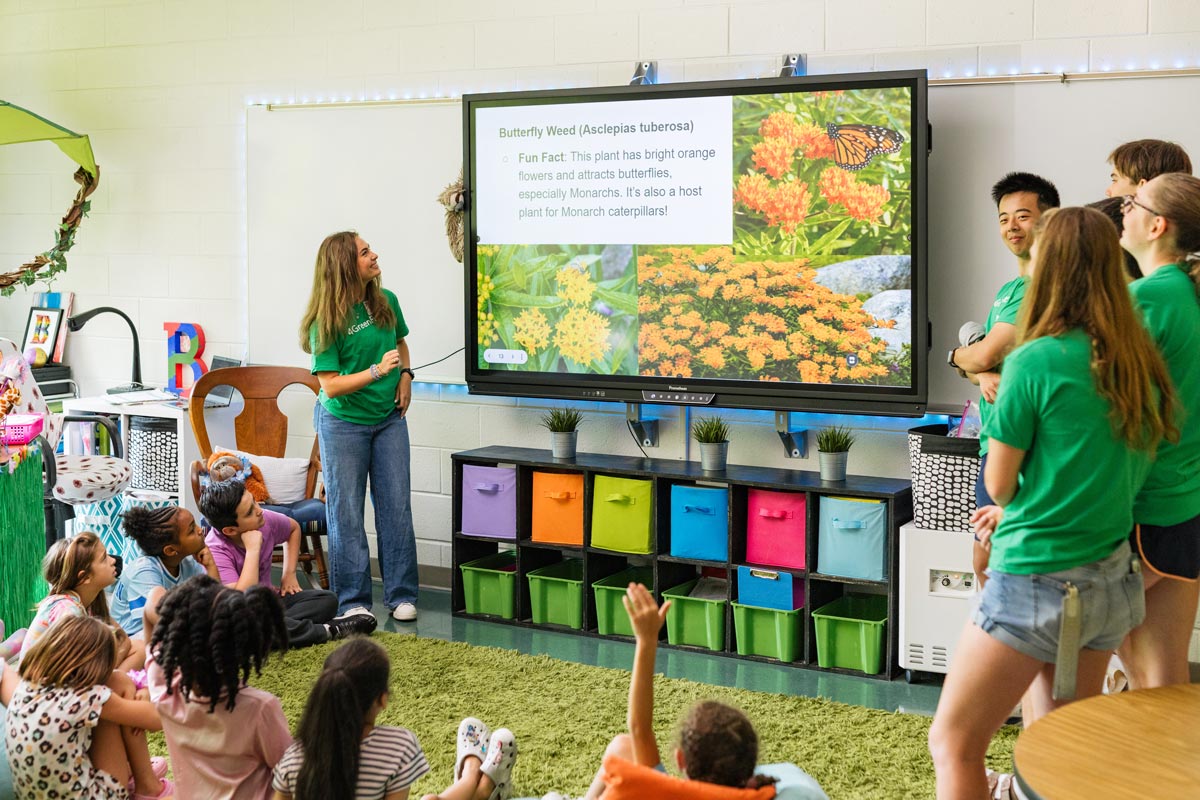 Children sit on a rug watching presenters in green shirts share a slide about Butterfly Weed and Monarch butterflies.