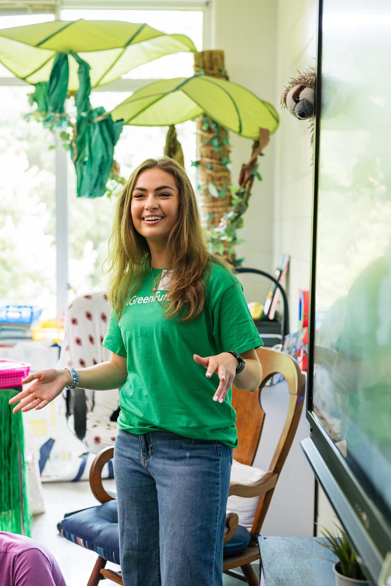 A person in a green shirt stands speaking in a classroom decorated with large green leaves and colorful materials.