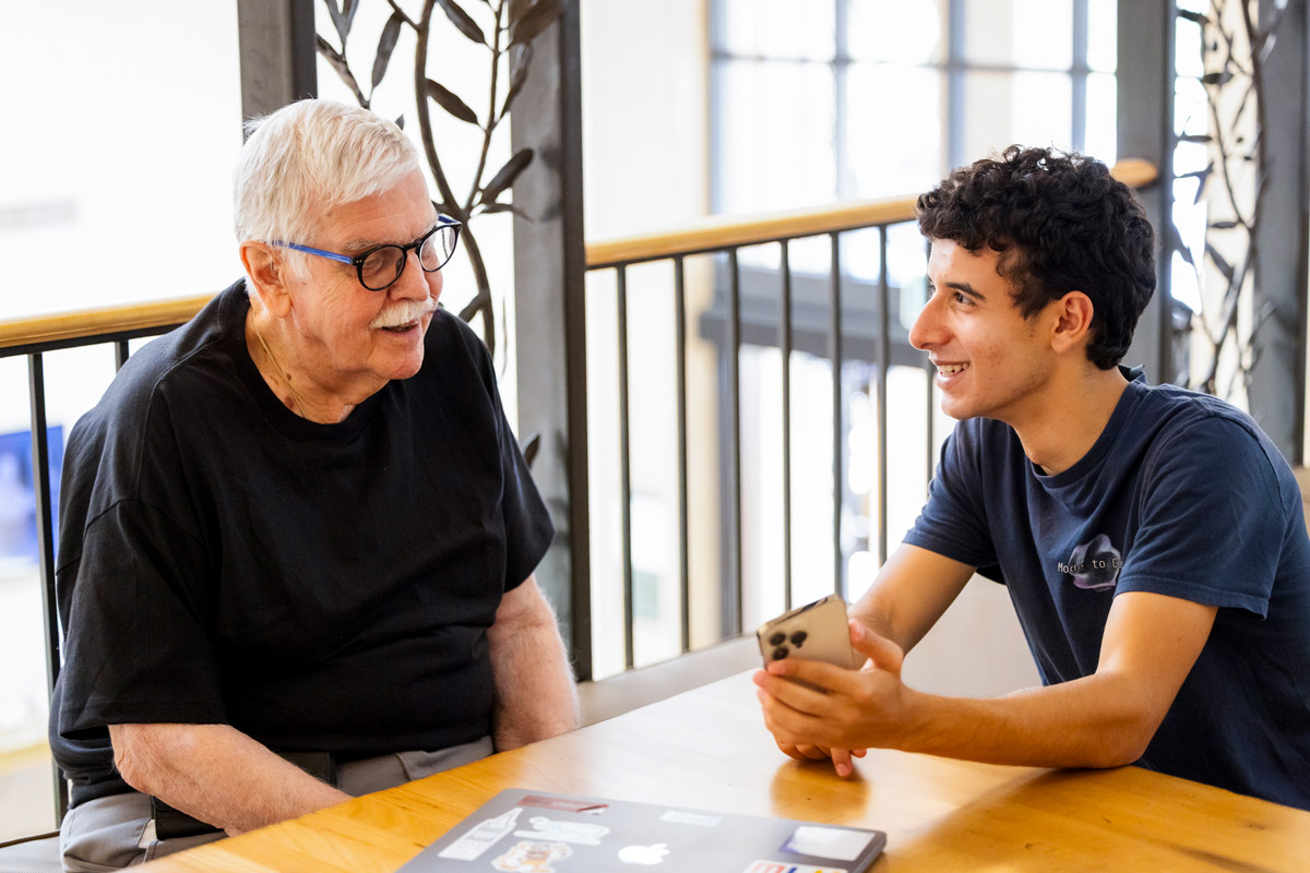 Two people sit at a wooden table talking as one holds a phone and a laptop with stickers rests between them.