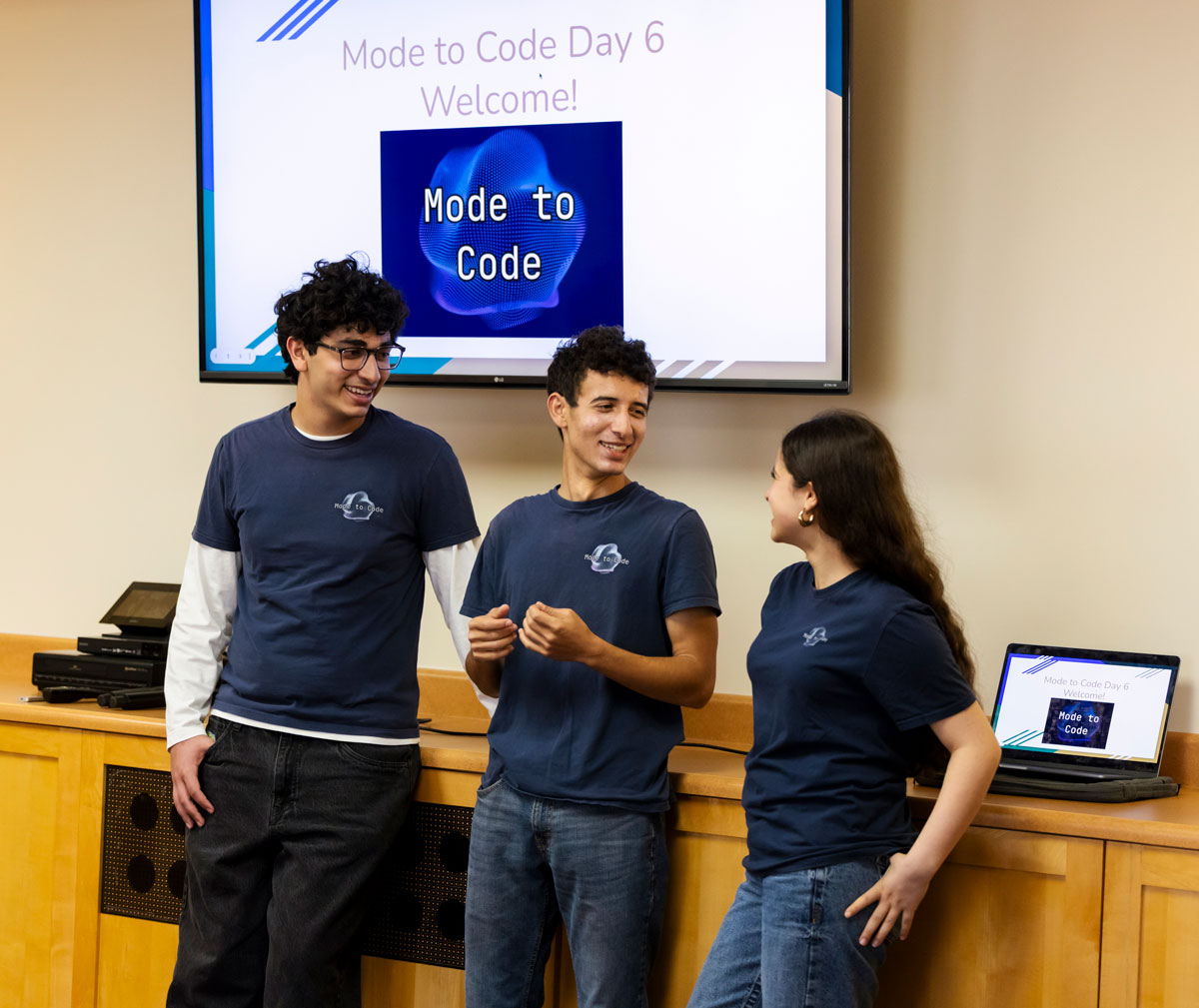 Three people stand presenting in front of a large screen displaying a “Mode to Code Day 6” welcome slide.